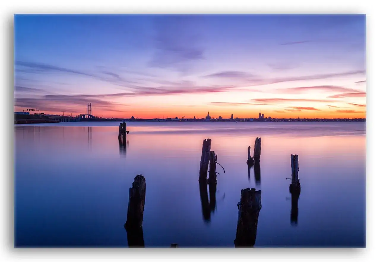 Sonnenuntergang &uuml;ber Stralsund: Alte Holzpf&auml;hle ragen aus dem ruhigen Wasser, im Hintergrund die Silhouette der Stadt und eine Br&uuml;cke. Ein Ort f&uuml;r Stralsund Geheimtipps.