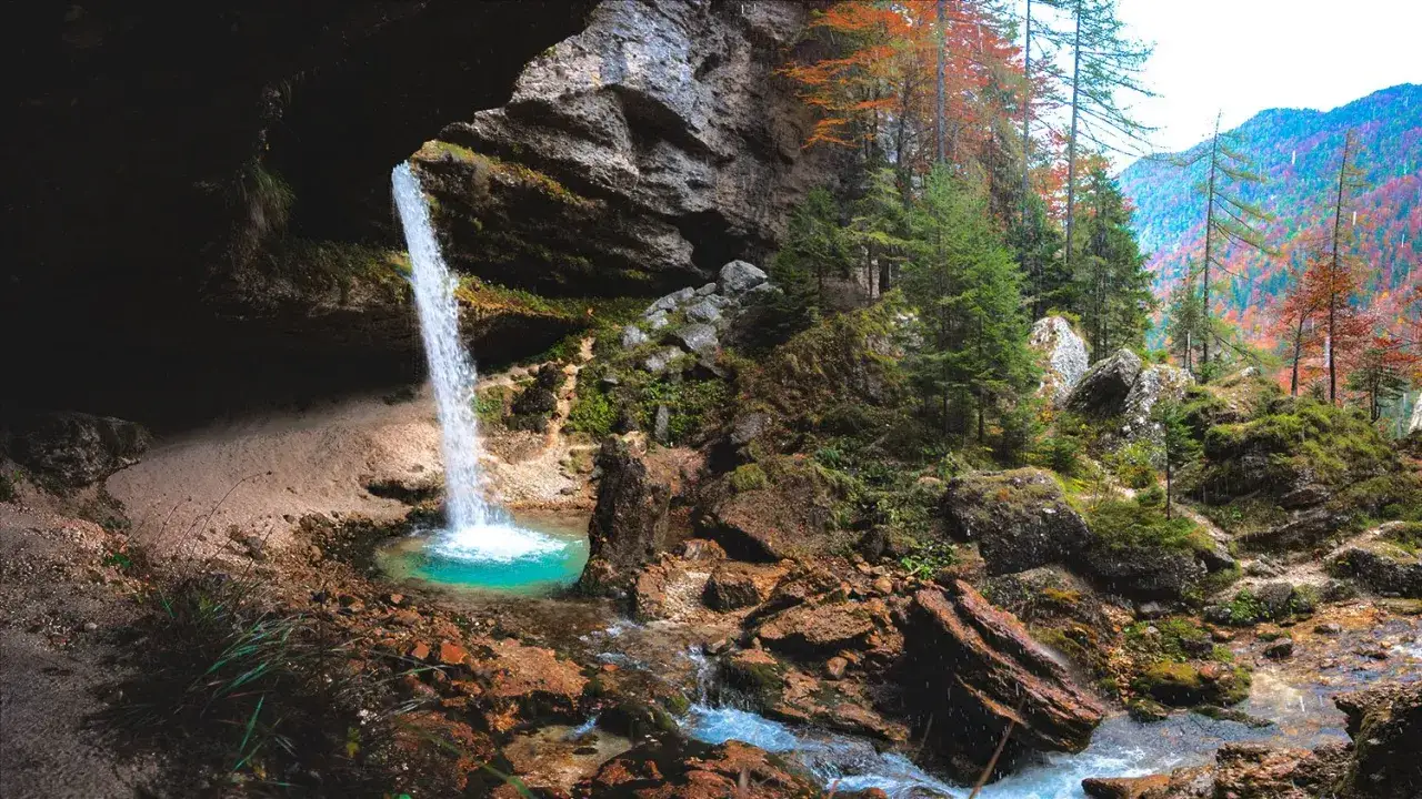 Ein Wasserfall stürzt in ein türkisfarbenes Becken im Vrata Tal. Herbstfarben schmücken die umliegenden Bäume.