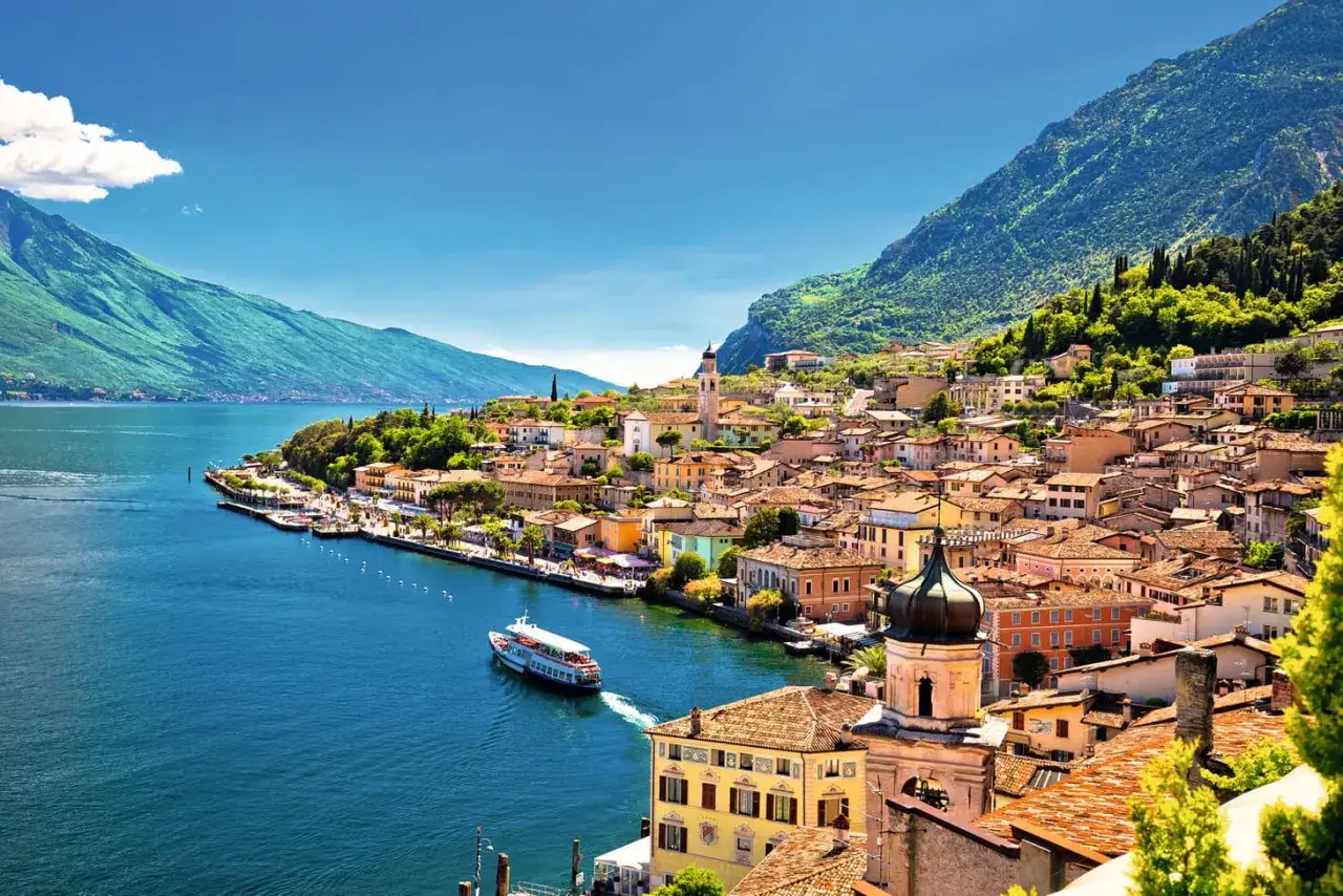 Bunte Häuser am Ufer des Gardasees, mit einem Schiff auf dem Wasser und grünen Bergen im Hintergrund. Ein malerischer Ort, der an den Lago Maggiore erinnert.