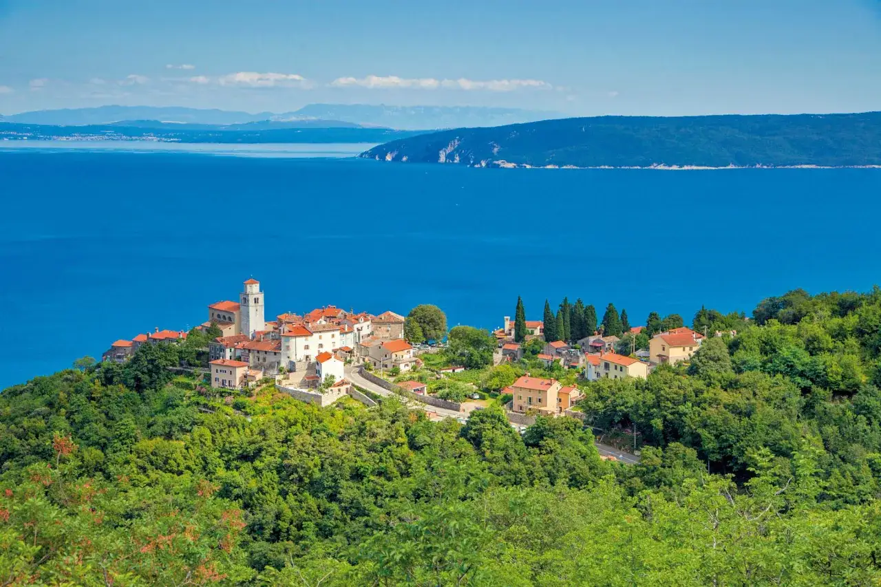 Ein malerisches Dorf mit roten Dächern und einem Kirchturm thront auf einem grünen Hügel über dem blauen Meer. Entdecken Sie die Geheimtipps von Opatija!