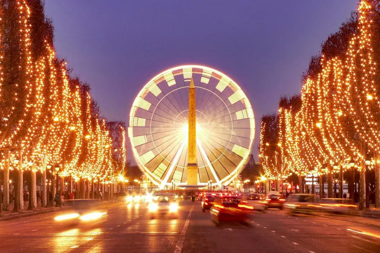 Paris Winter: Ein Riesenrad und ein Obelisk im Lichterglanz, Bäume mit Lichterketten gesäumt, Autos fahren vorbei.