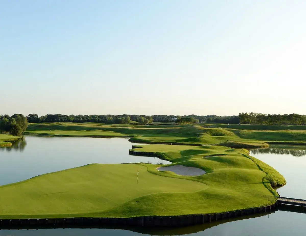 Grüne Fairways und Wasserhindernisse auf dem Le Golf National. Ein Steg führt zu einer Insel mit Bunker und Fahne.