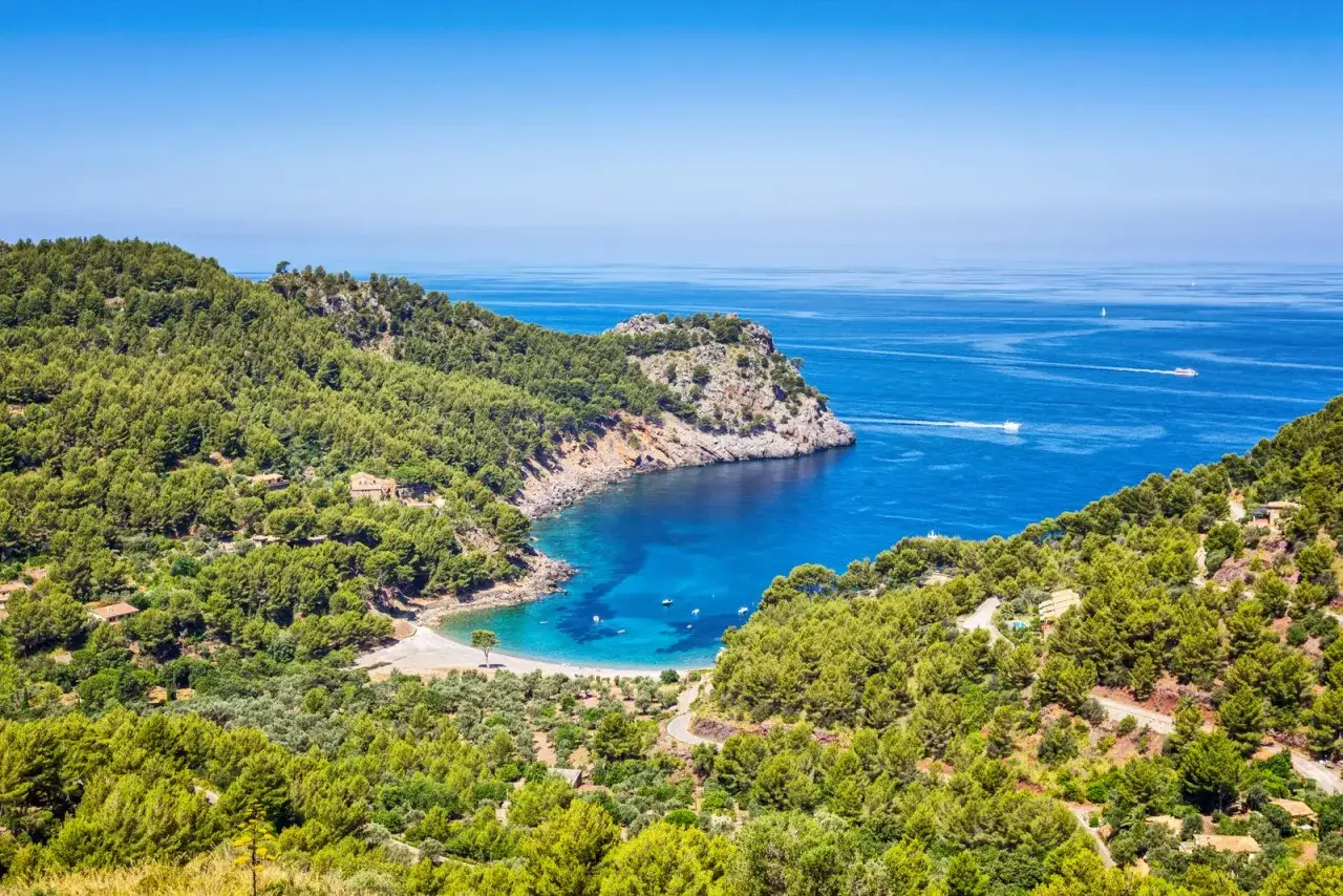 Traumhafte Bucht mit türkisfarbenem Wasser, umgeben von grünen Hügeln des Gebirge Mallorca. Boote gleiten auf dem Meer.