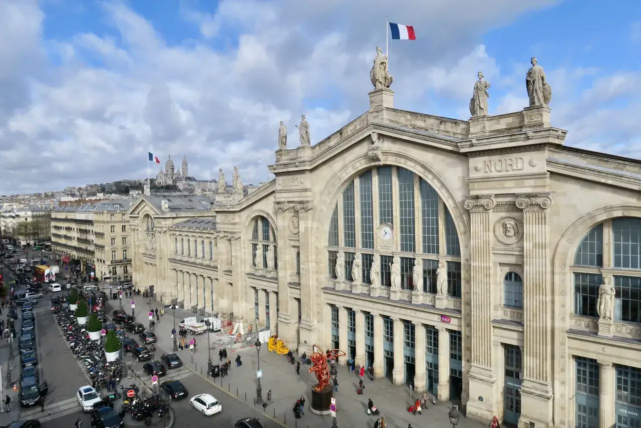 Der Gare du Nord in Paris, ein imposanter Bahnhof mit Skulpturen und der französischen Flagge, im Hintergrund die Sacré-Cœur-Basilika.