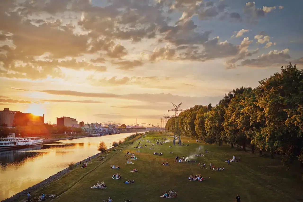 Sonnenuntergang an der Neckarwiese Mannheim. Menschen entspannen auf der Wiese, w&auml;hrend ein Schiff auf dem Neckar vorbeif&auml;hrt.