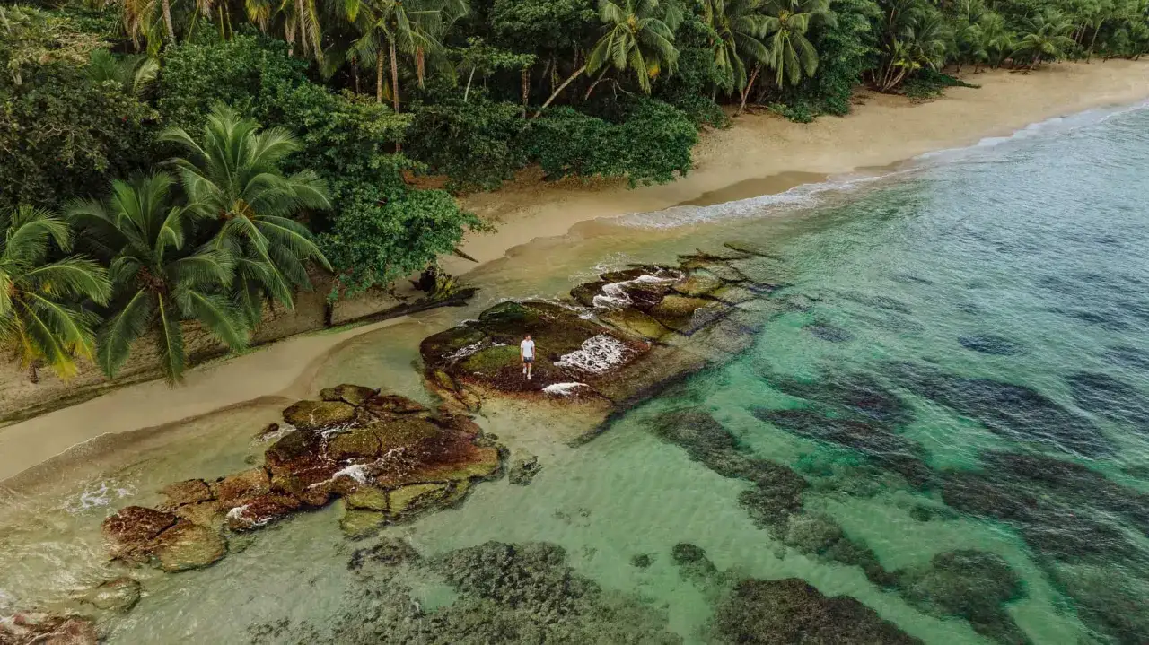 Mann steht auf Felsen am puerto viejo strand, umgeben von &uuml;ppiger Vegetation und t&uuml;rkisfarbenem Wasser.