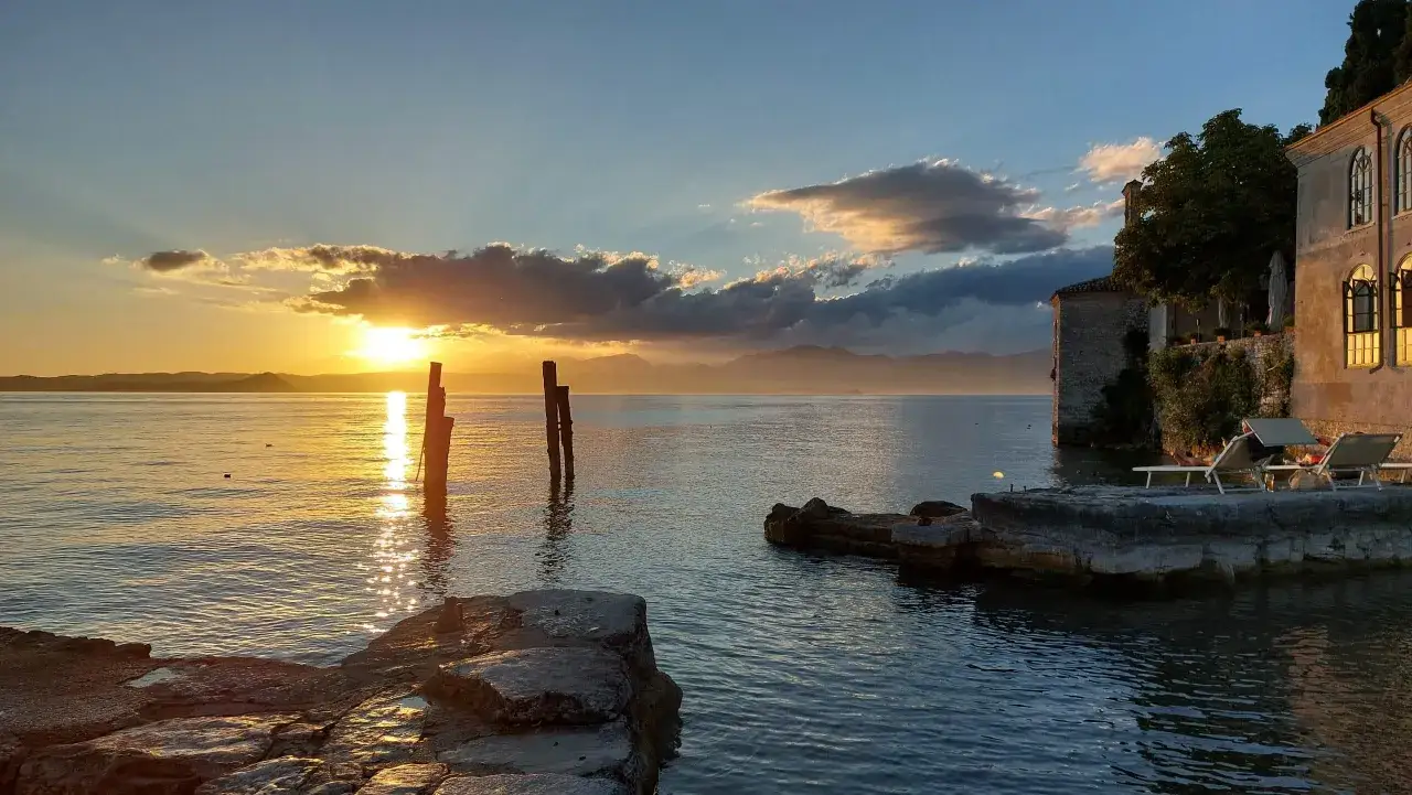 Sonnenuntergang am Punta San Vigilio. Goldene Strahlen spiegeln sich im ruhigen Wasser. Ein Gebäude mit Bögen und Liegestühle am Ufer.