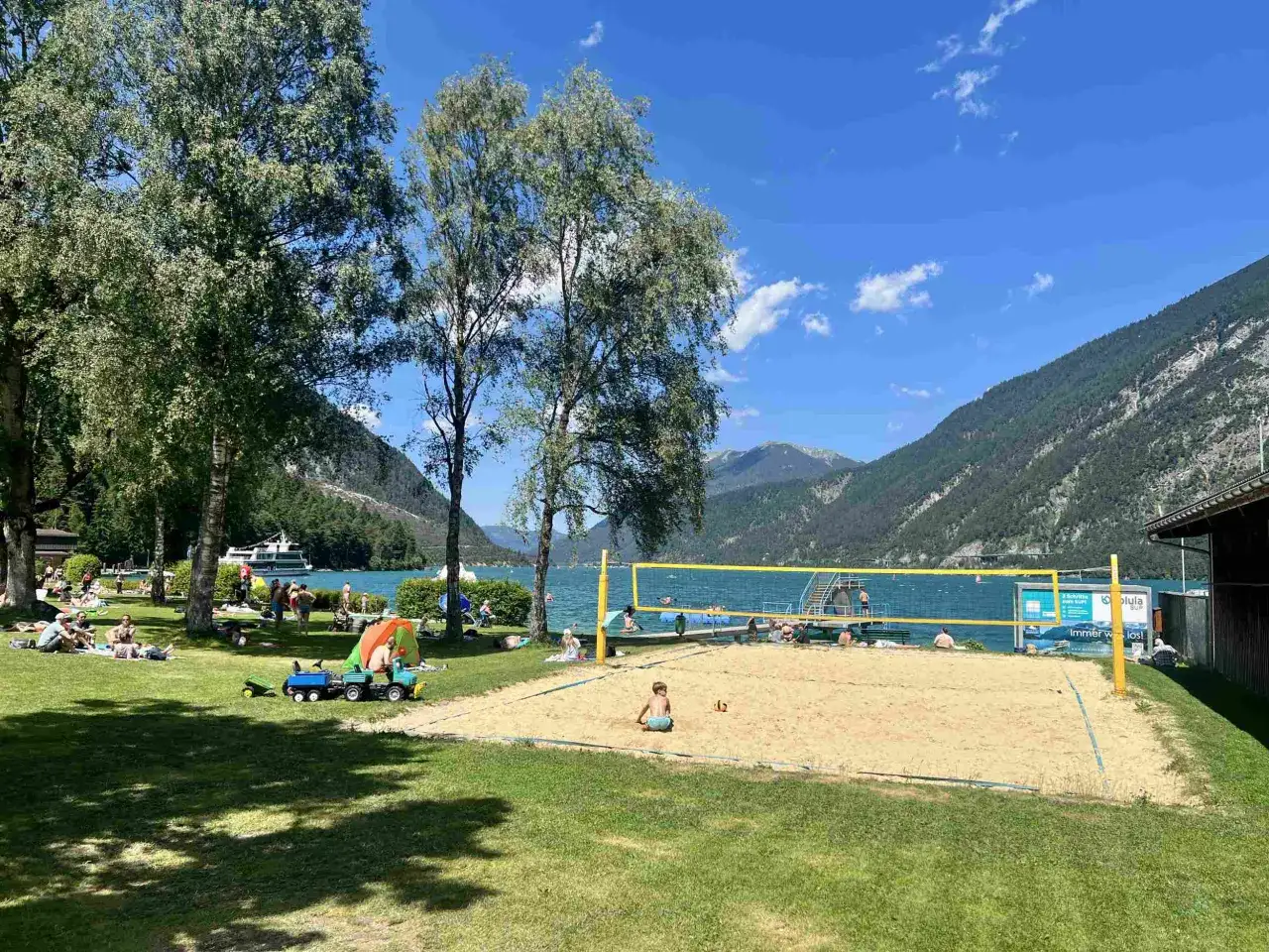 Sommerliche Szene am Achensee mit Bergen im Hintergrund. Menschen entspannen am Ufer, spielen Beachvolleyball und genie&szlig;en die Sonne.
