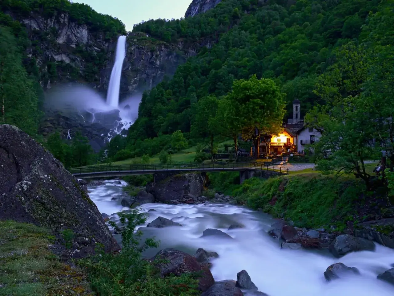 Ein einsames Haus mit Licht am Wasserfall, ein Schweiz wandern Geheimtipp. Ein Fluss flie&szlig;t vorbei, umgeben von gr&uuml;nen W&auml;ldern.