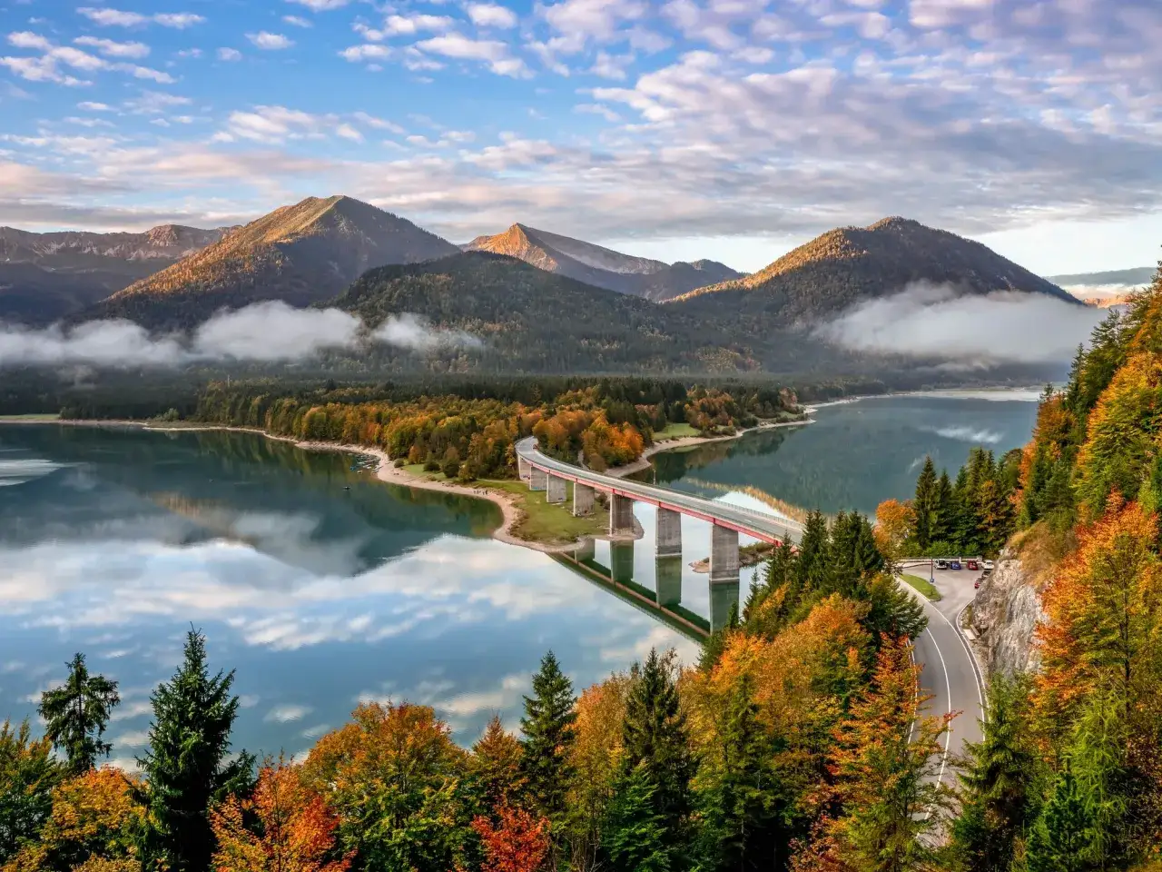 Herbstliche Farben umgeben einen See mit einer Br&uuml;cke, ein wahres Naturwunder Deutschland. Berge und Wolken spiegeln sich im ruhigen Wasser.