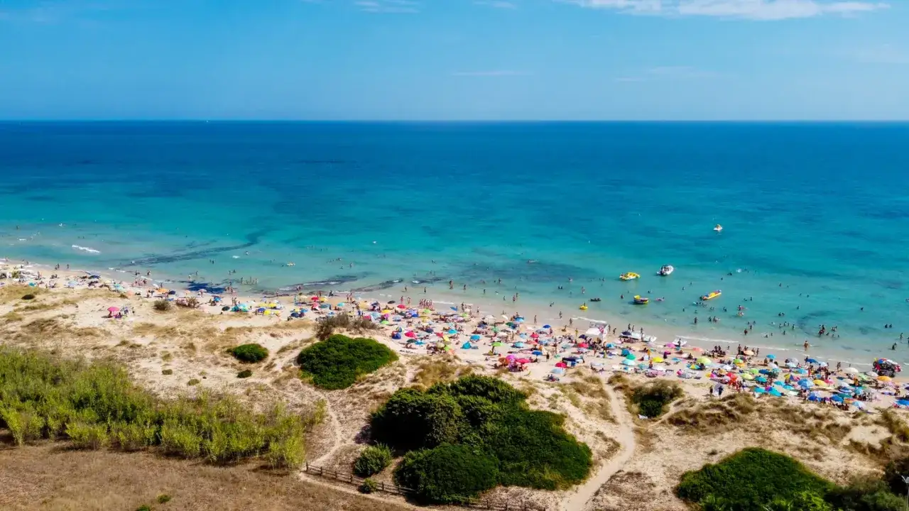 Sonniger Strand in Apulien, Italien, mit vielen bunten Sonnenschirmen und Menschen, die im türkisfarbenen Meer schwimmen.