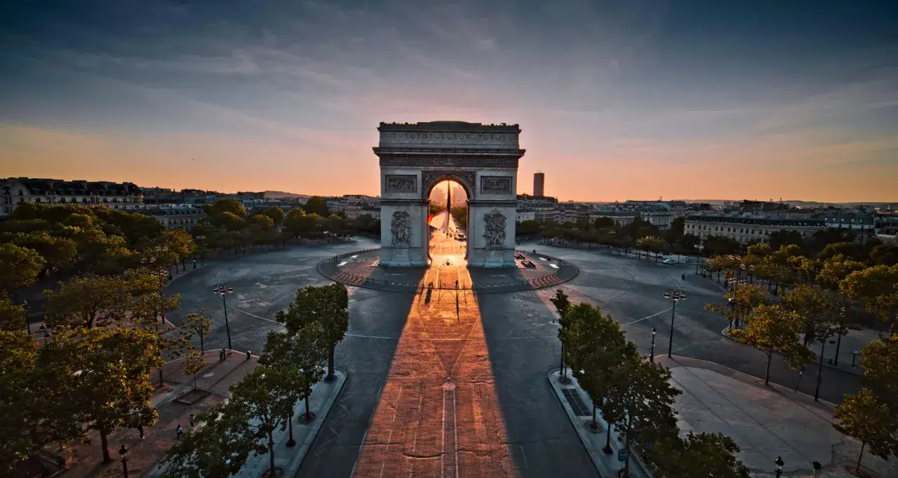 Der Arc de Triomphe in Paris im goldenen Licht der Morgensonne. Ein langer Schatten fällt über den Platz.