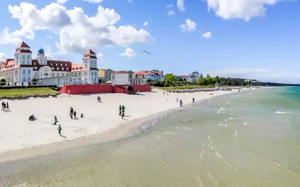 Ein sonniger Ostsee Strand mit Menschen, die spazieren gehen und Kinder spielen. Im Hintergrund ein prächtiges Gebäude im Bäderstil.