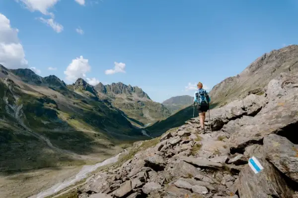 Frau mit Rucksack und Stöcken wandert auf felsigem Pfad im Montafon. Bergpanorama mit Tälern und Gipfeln.
