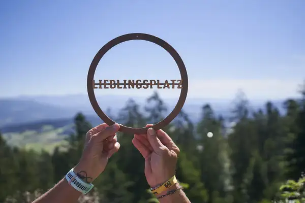 Hände halten ein Schild mit "LIEBLINGSPLATZ" vor einer malerischen Bergkulisse. Ein echter Geheimtipp im Bayerischen Wald.