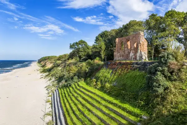 Ruinen einer alten Kirche thronen über einem Strand an der polnischen Ostsee. Terrassierte grüne Hänge führen zum Meer.