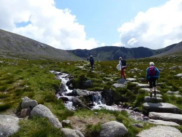 Drei Wanderer genießen das Cairngorms National Park Wandern entlang eines steinigen Pfades mit einem plätschernden Bach.