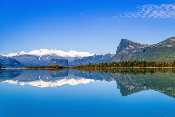 Spiegelglatte Wasserfläche reflektiert schneebedeckte Berge und herbstliche Bäume in einem schwedischen Nationalpark.
