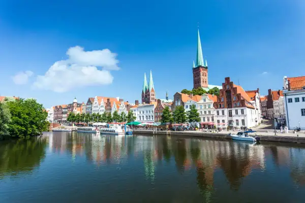 Historische Gebäude und Kirchtürme säumen das Wasser in Lübeck, einem der schönsten Ostsee Orte. Boote liegen am Kai, die Sonne scheint.