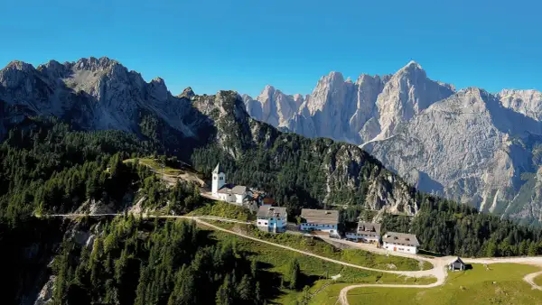 Alpe-Adria-Trail Erfahrungsbericht: Malerisches Bergdorf mit Kirche und Häusern, umgeben von Nadelwäldern und imposanten Gipfeln.