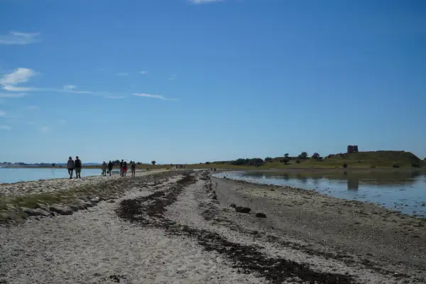 Menschen spazieren auf einem sandigen Weg entlang des Wassers in Richtung der Mols Bjerge mit einer alten Burgruine auf dem Hügel.