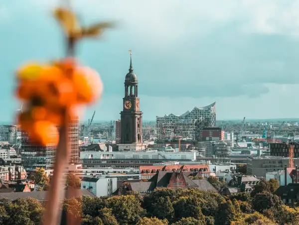 Blick auf Hamburg sehenswertes: Kirchturm St. Michaelis und Elbphilharmonie vor bewölktem Himmel. Im Vordergrund unscharfe orangefarbene Blüten.