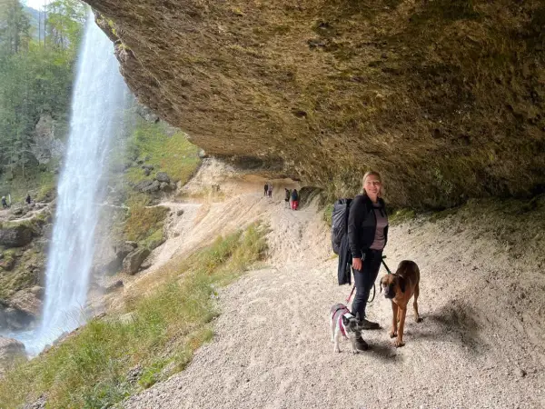 Frau mit zwei Hunden unter einem Felsvorsprung, während ein Wasserfall in der Nähe herabstürzt. Ein perfekter Ort, um die Natur zu genießen.