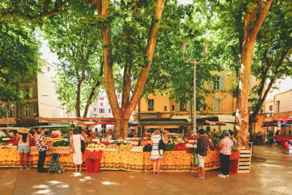 Bunter Markt in Aix-en-Provence unter schattigen Bäumen. Menschen kaufen frisches Obst und Gemüse an Ständen mit provenzalischen Stoffen.