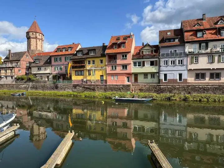 Bunte Häuserfront am Flussufer, ein Turm ragt empor. Hier könnte das Taubertal sein, mit Spiegelungen im Wasser.